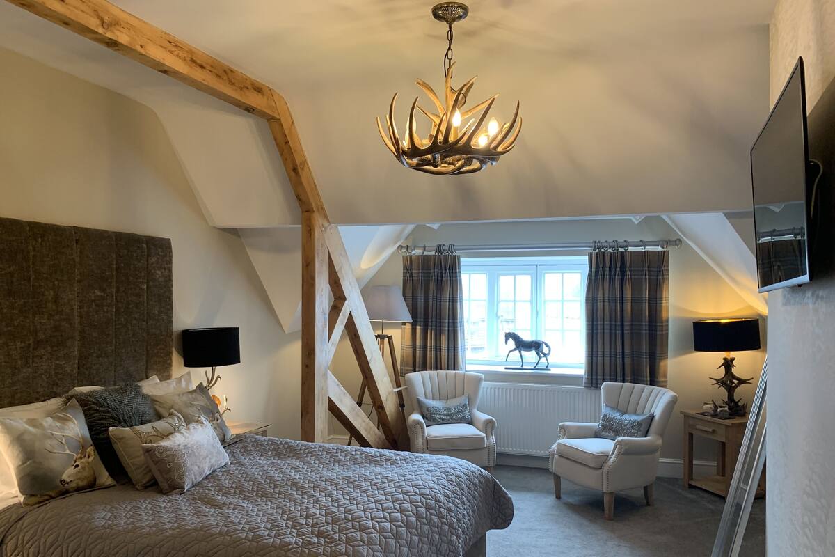 A bedroom with a large bed, two armchairs, a window with curtains, a chandelier, and a mounted TV.