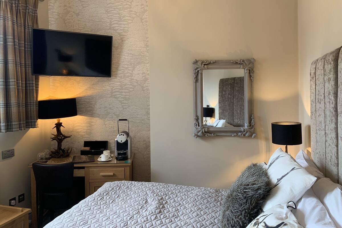 A hotel room with a bed, a desk with a computer, a flat-screen TV, a coffee machine, and a mirror.