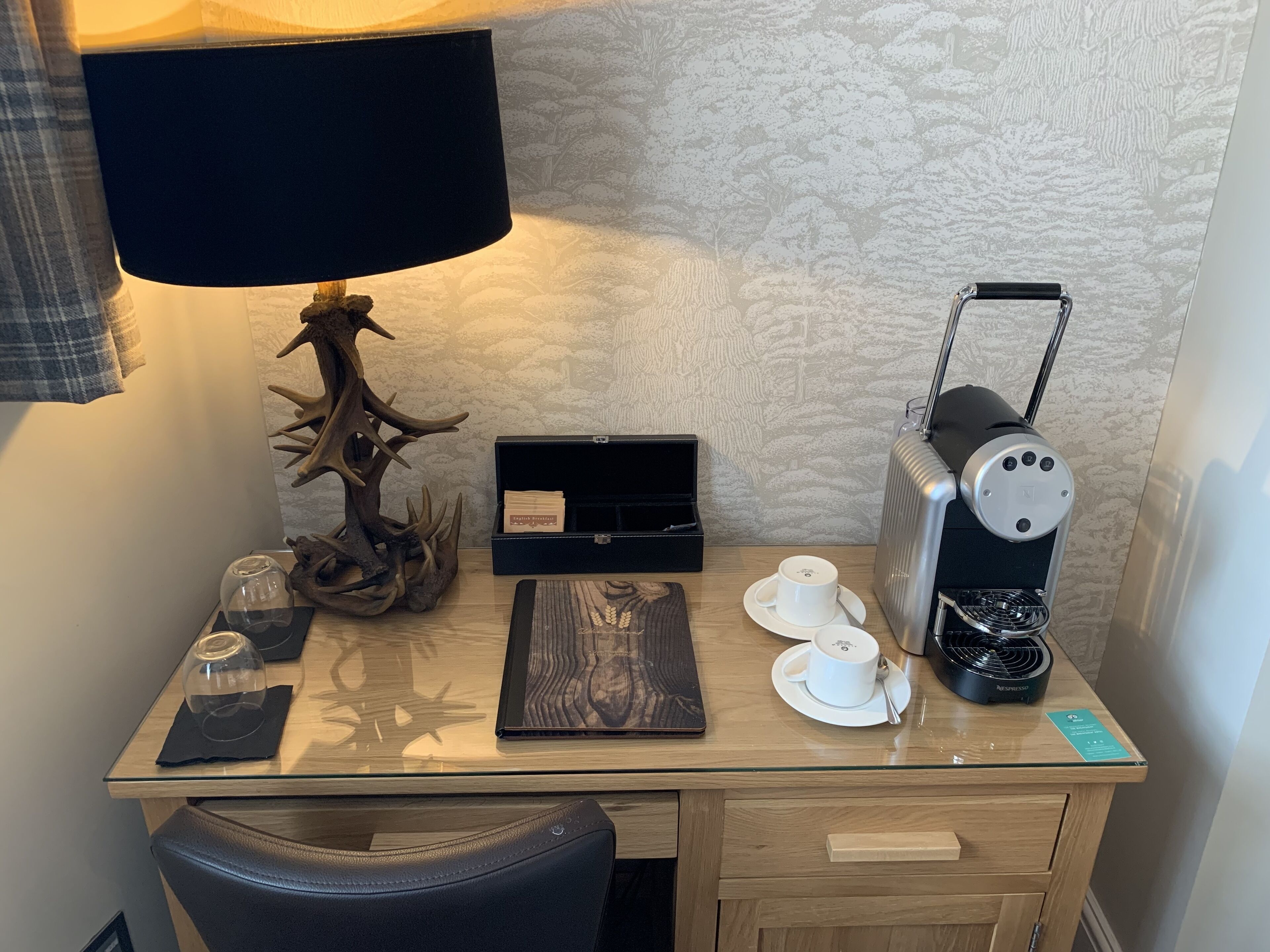Un escritorio de hotel con una lámpara, una cafetera, un vaso de agua, una taza y una caja de pañuelos.