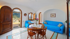 A room with a blue sofa, wooden chairs, a round table with a vase of flowers, and a view of the mountains through an arched doorway. - Melodia (Praiano)