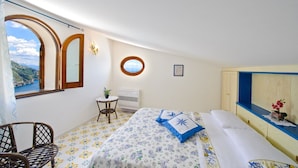 A bedroom with a bed, a chair, a small table, a window, and a wall-mounted air conditioner. - Melodia (Praiano)