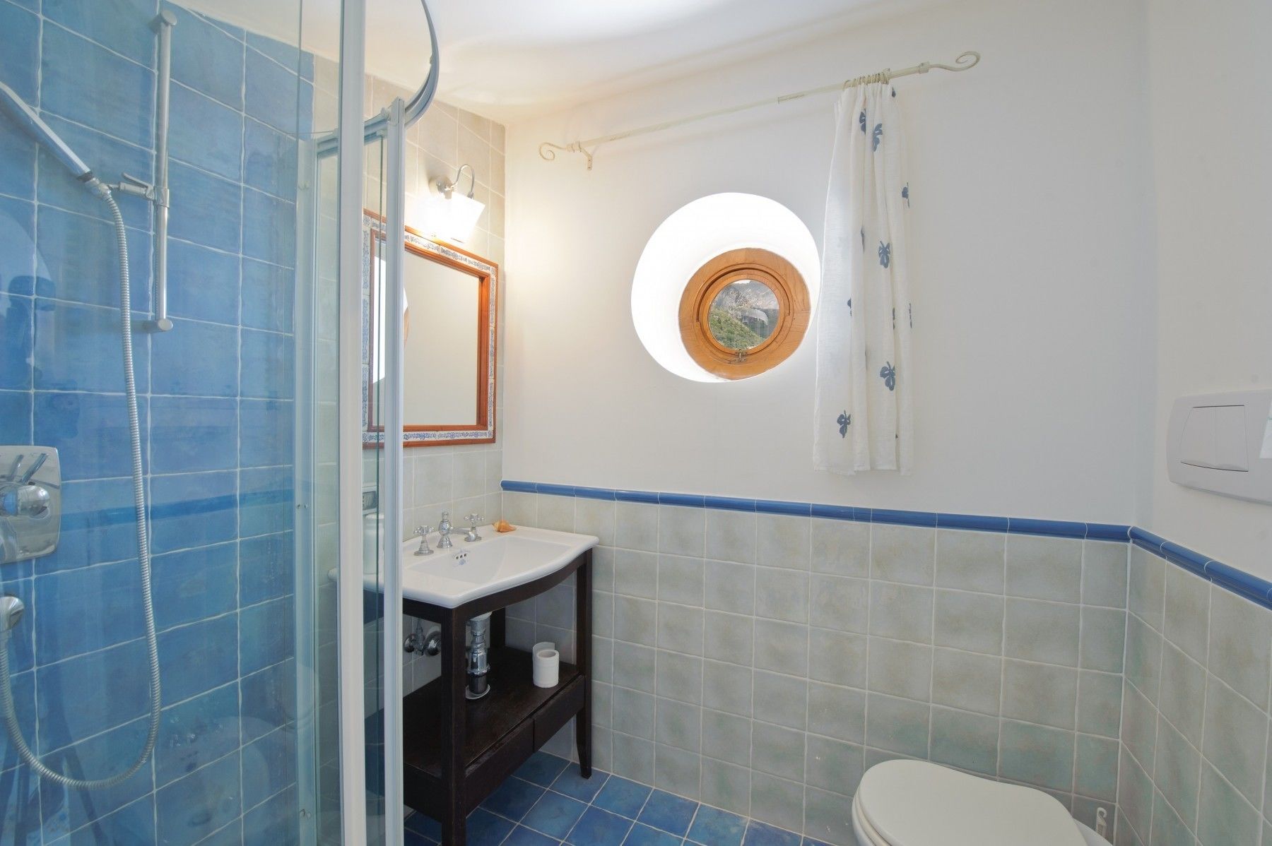 A bathroom with a glass-enclosed shower, a sink with a mirror, a toilet, and a small window with a wooden frame.