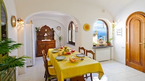 A dining area with a table set for four, a wooden cabinet, and a view of the sea through an arched window. - Melodia (Praiano)