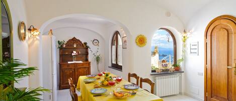 A dining area with a table set for four, a wooden cabinet, and a view of the sea through an arched window.