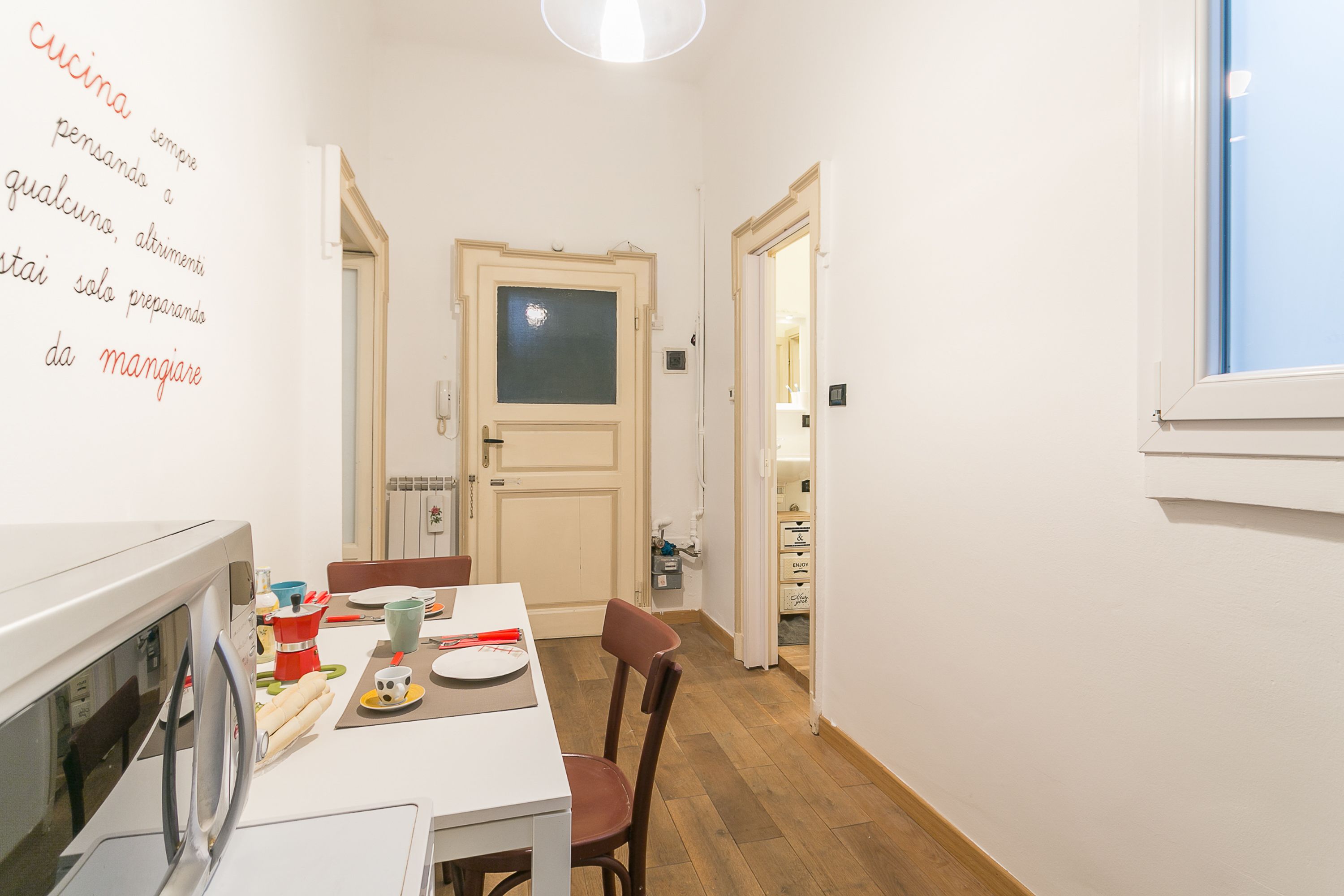 Eine schmale Küche mit Esstisch, Stühlen und einer Mikrowelle. Rechts ist ein Fenster und eine Tür führt in einen anderen Raum.
