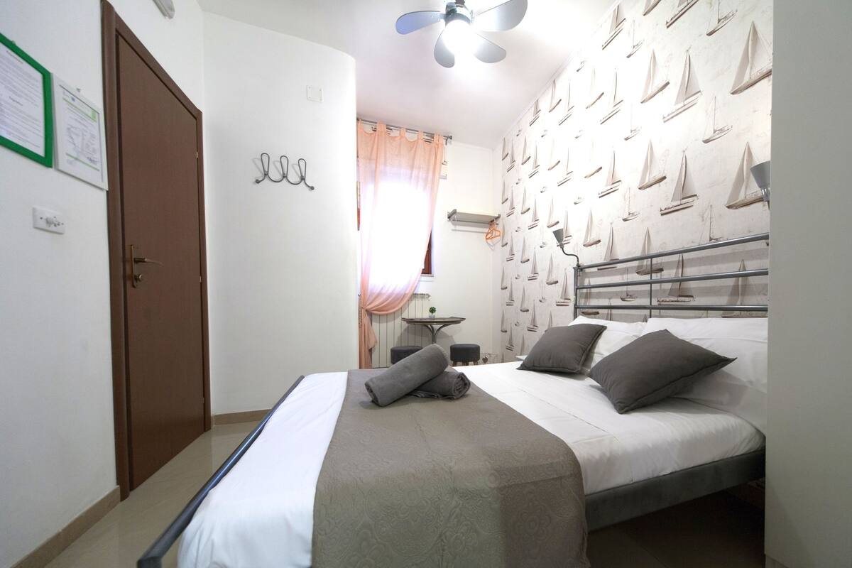 A bedroom with a bed, a ceiling fan, a wall with sailboat decorations, a door, and a small table with a chair.