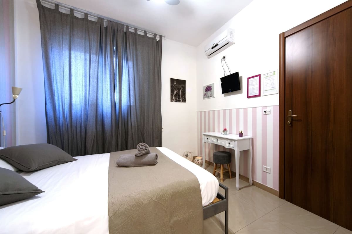 A bedroom with a bed, a desk, a chair, a wardrobe, and an air conditioning unit.