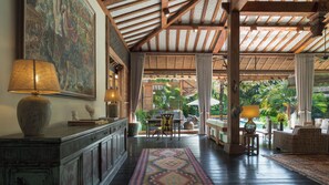 A spacious room with a high ceiling, wooden beams, a large painting, a dining area with a table and chairs, and a view of lush greenery outside.