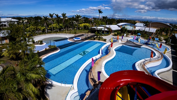 A large swimming pool with multiple lanes, surrounded by palm trees and a red slide.