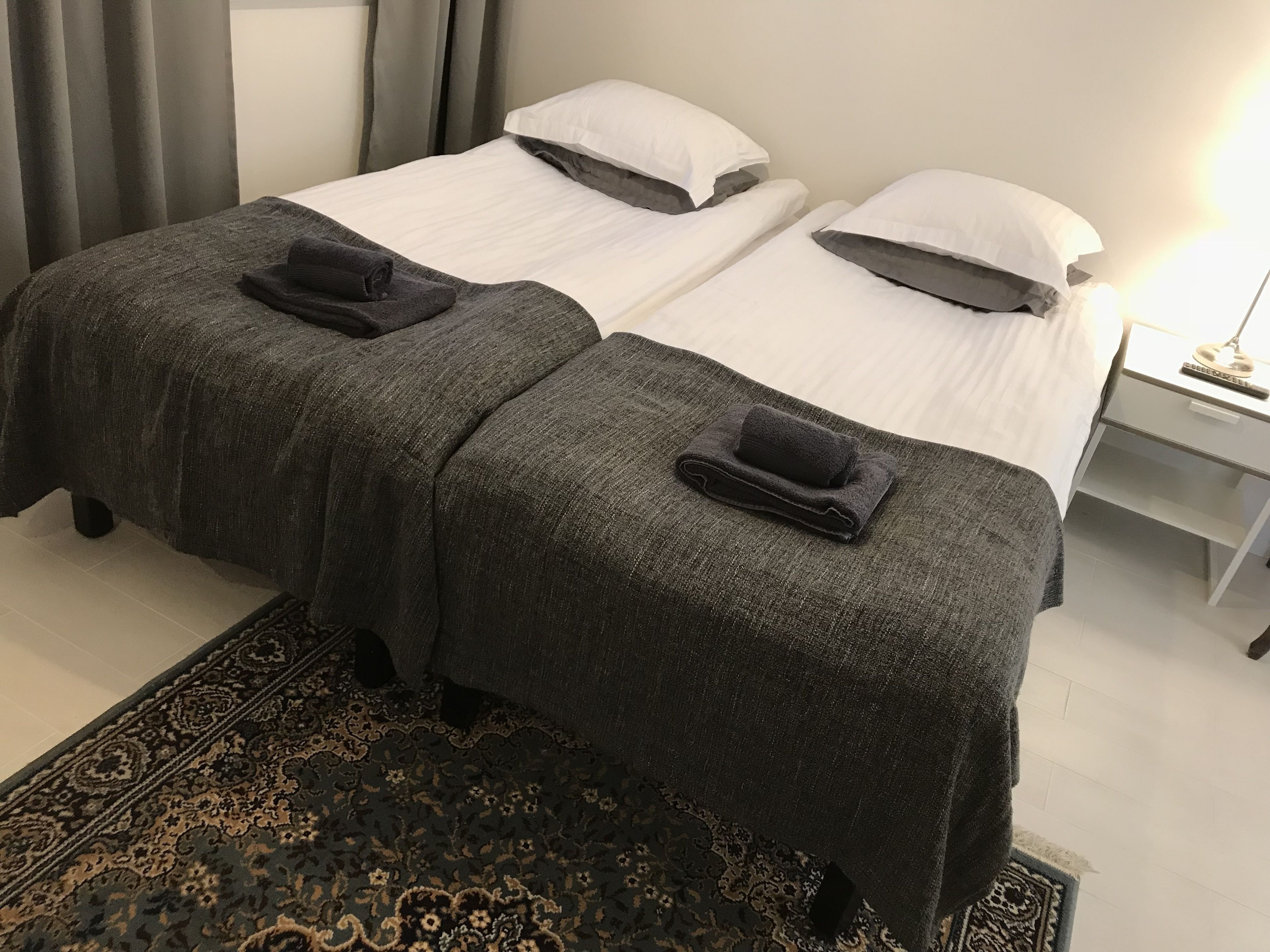 A hotel room with two beds, each with a dark grey blanket and a matching towel. There is a nightstand with a lamp and a small drawer.