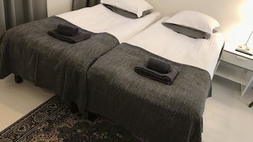 A hotel room with two beds, each with a dark grey blanket and a matching towel. There is a nightstand with a lamp and a small drawer.