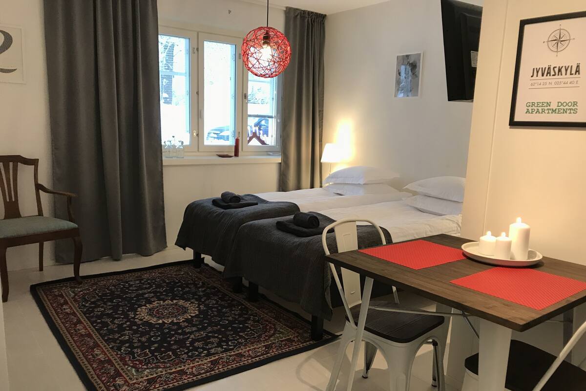 A bedroom with two beds, a window, a desk with chairs, a red rug, and a wall-mounted light fixture.