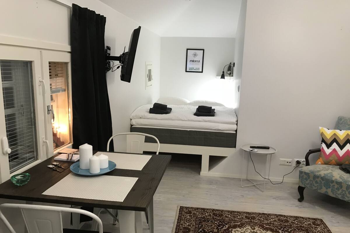 A small, modern room with a bed, a desk, and a chair. There is a window with curtains, a framed picture on the wall, and a rug on the floor.
