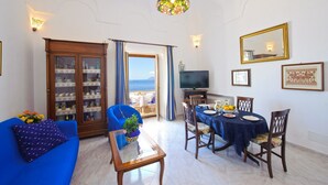 Un salon avec un canapé bleu, une table basse en verre, une table à manger avec des chaises, un téléviseur et une vue sur la mer par une porte.