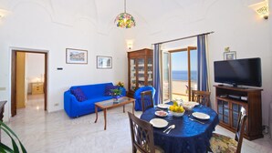 Un salon avec un canapé bleu, une table à manger dressée avec des assiettes et des verres, un téléviseur et une vue sur la mer par une porte vitrée.