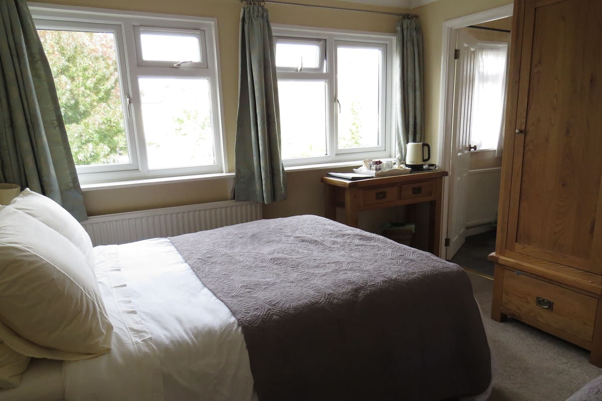 A bedroom with a bed, a wooden wardrobe, a desk with a kettle, and a window with curtains.