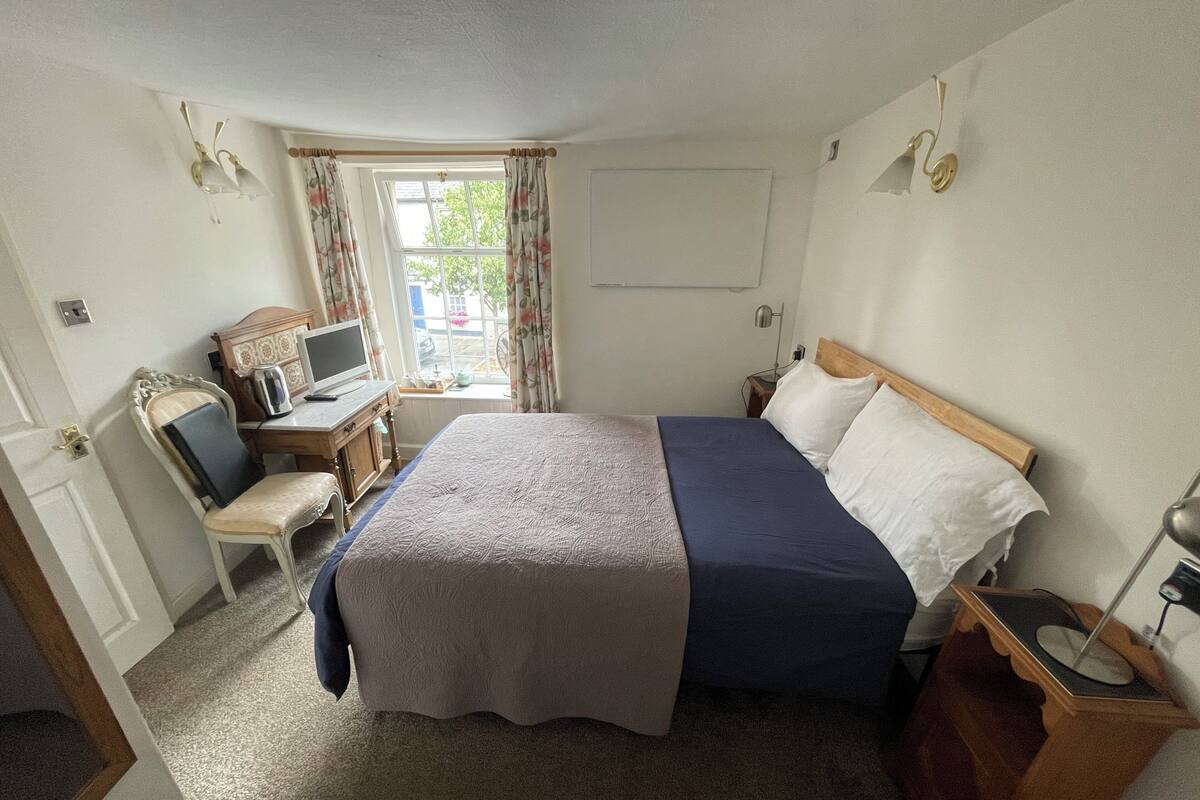 A hotel room with a bed, a desk with a computer, a chair, a window with curtains, and a wall-mounted lamp.