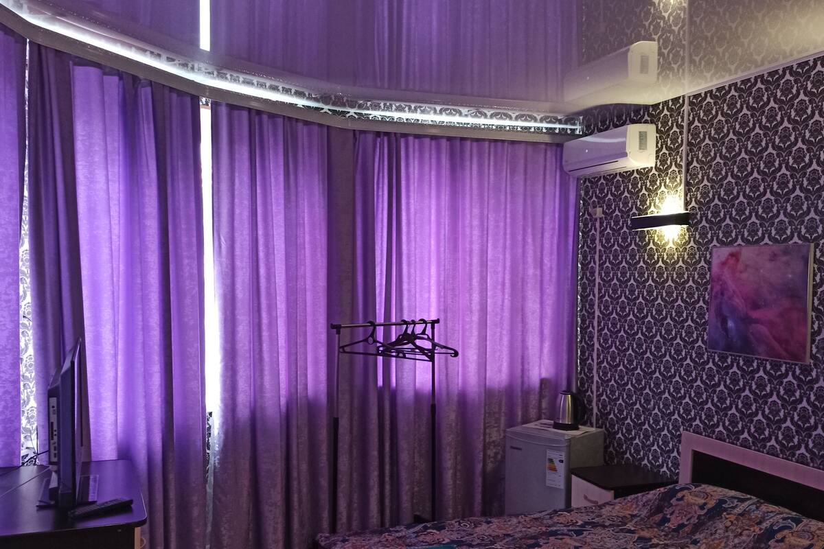 Ein Schlafzimmer mit violetten Vorhängen, einem Bett mit gemustertem Überwurf, einem Nachttisch, einer Lampe und einer Wand-Klimaanlage.