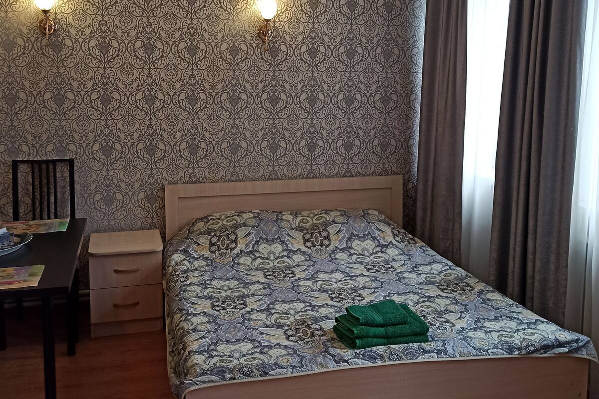 Ein ordentlich eingerichtetes Schlafzimmer mit einem gemusterten Bettbezug, einem Nachttisch mit Lampe und einem Sessel.
