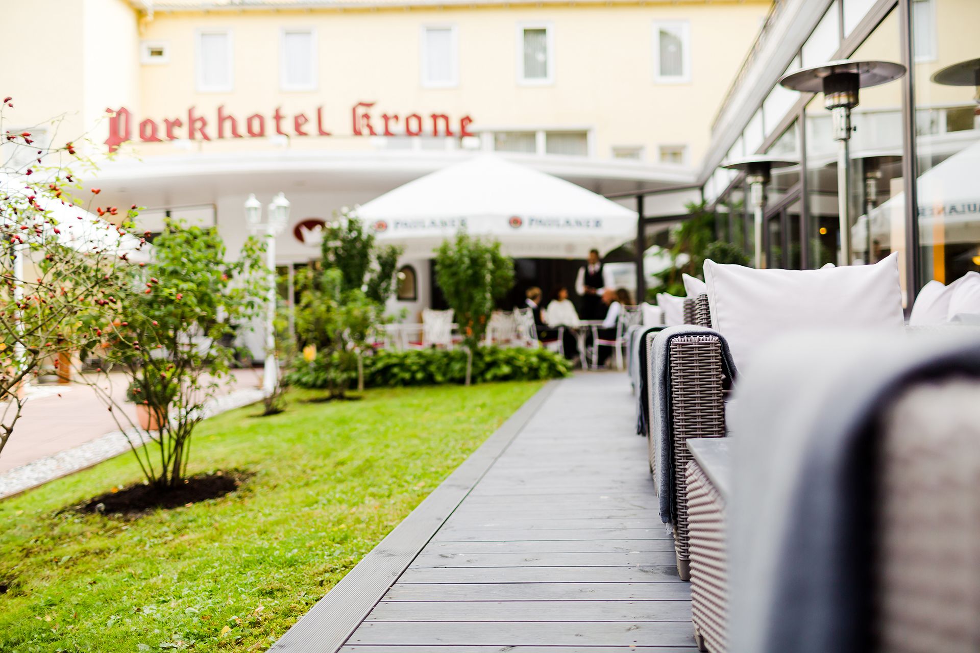 A pathway leading to outdoor seating with umbrellas, a sign for Parkhotel Krone, and a building with large windows.