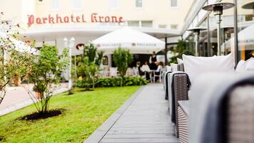 A pathway leading to outdoor seating with umbrellas, a sign for Parkhotel Krone, and a building with large windows.