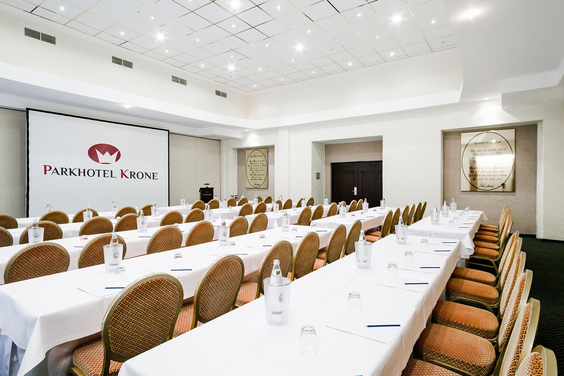 Een congreszaal in het Parkhotel Krone met meerdere tafels, stoelen en een presentatiescherm.