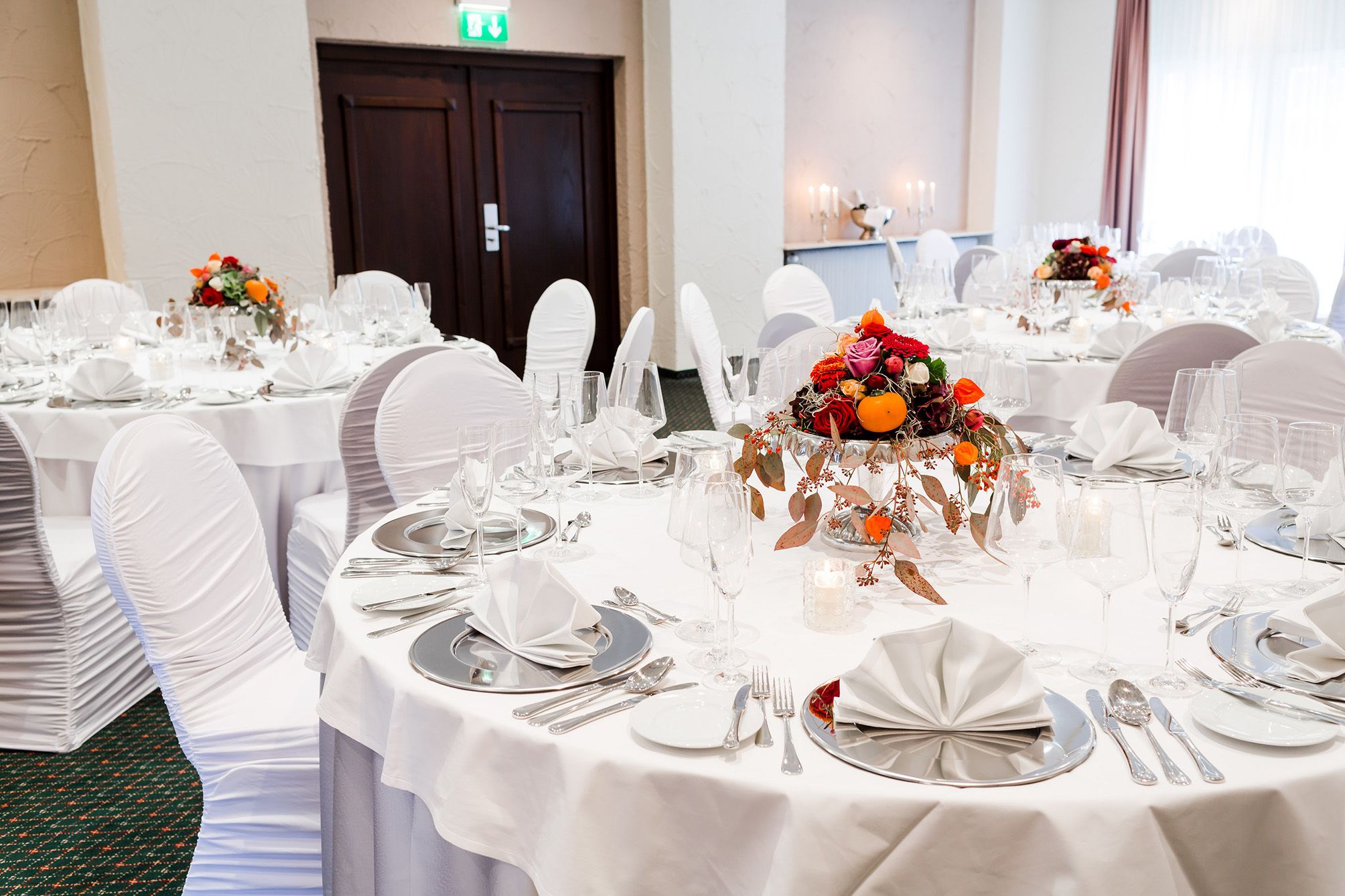 Ein Bankettsaal mit runden Tischen, festlich gedeckt für eine formelle Veranstaltung, mit weißen Tischdecken, Besteck und floralen Mittelpunkten.