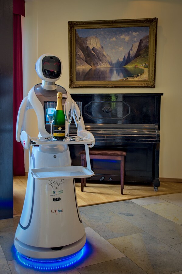 Un robot, avec une bouteille de champagne et des verres sur son plateau, un piano et un tableau encadré accroché au mur.