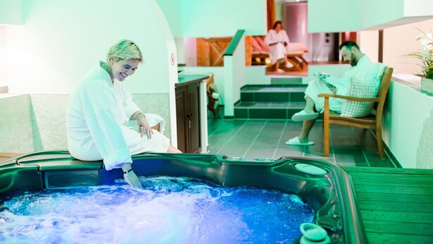 Et spa-område med jacuzzi, en person i badekåbe og en anden person, der læser en bog.