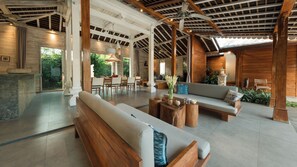 A spacious indoor area with a wooden ceiling, a sofa set, a dining table, and a seating area with a view of greenery outside. - Mannao Estate by Villa Finder (Kerobokan)