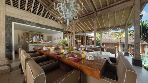 A spacious dining area with a high ceiling and a large chandelier, featuring a long wooden table set for a meal.