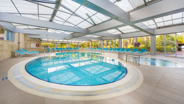 Una amplia piscina cubierta con varias pistas, rodeada de sillones azules y un techo de cristal que deja entrar luz natural.