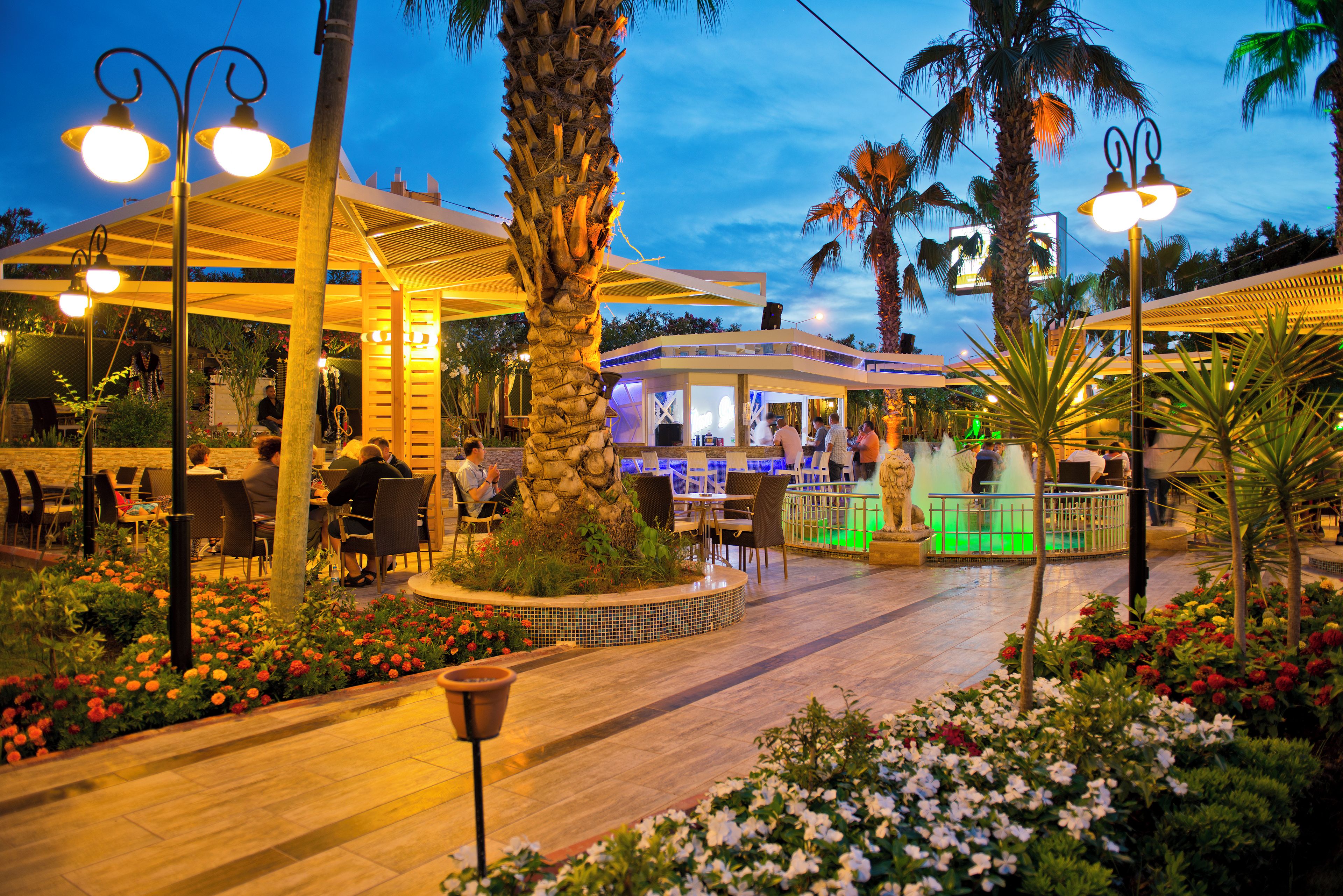 An outdoor restaurant area with tables and chairs, surrounded by palm trees and flower beds.