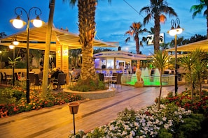 An outdoor restaurant area with tables and chairs, surrounded by palm trees and flower beds. - Beach Club Doganay (Alanya)