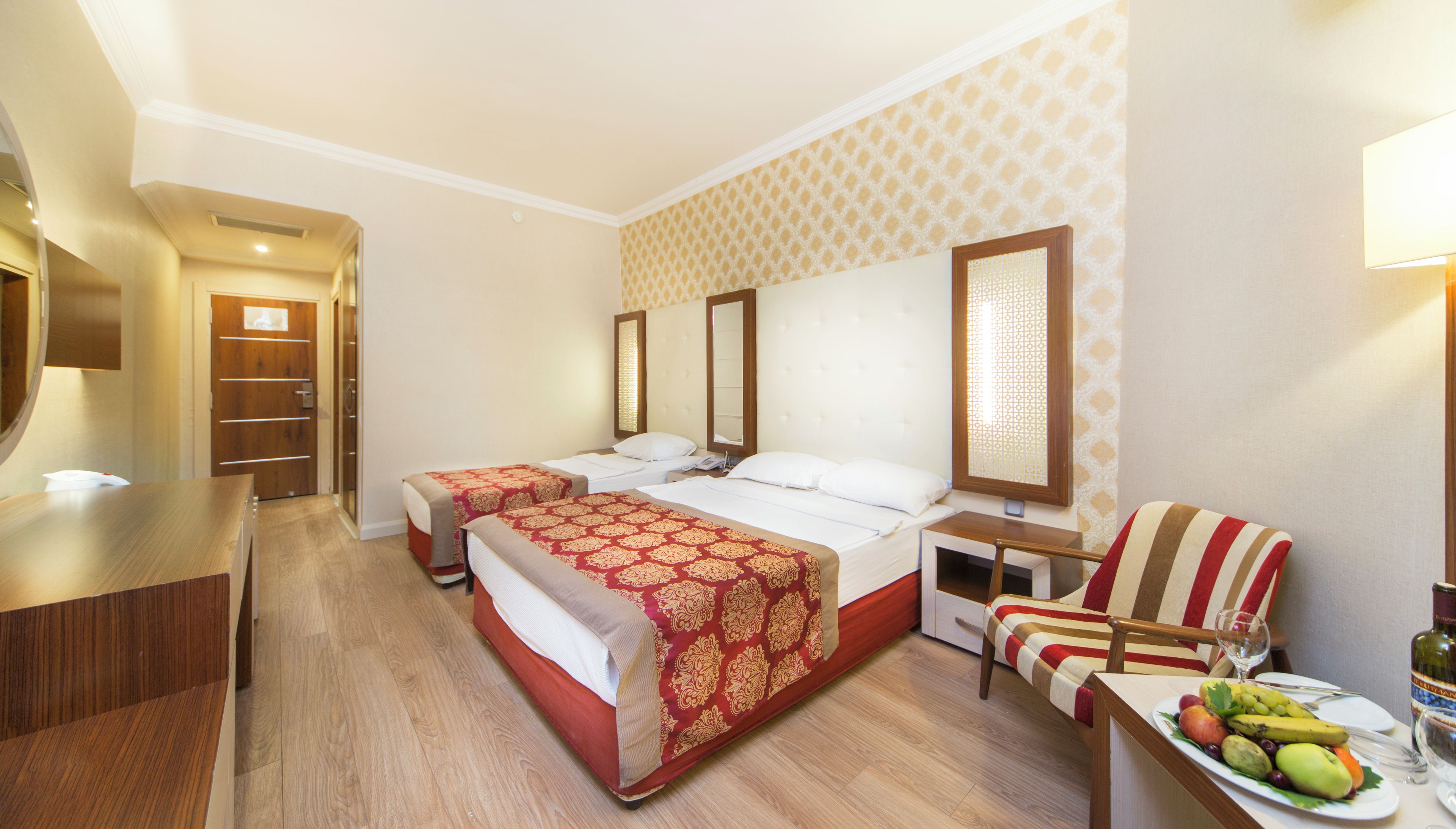A hotel room with two beds, a desk, a chair, and a dining table with fruit and a bottle.