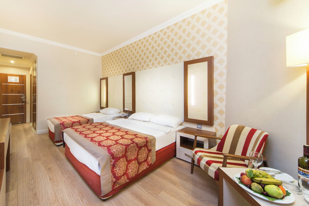Une chambre d’hôtel avec deux lits, un bureau, une chaise et une table à manger avec des fruits et une bouteille.