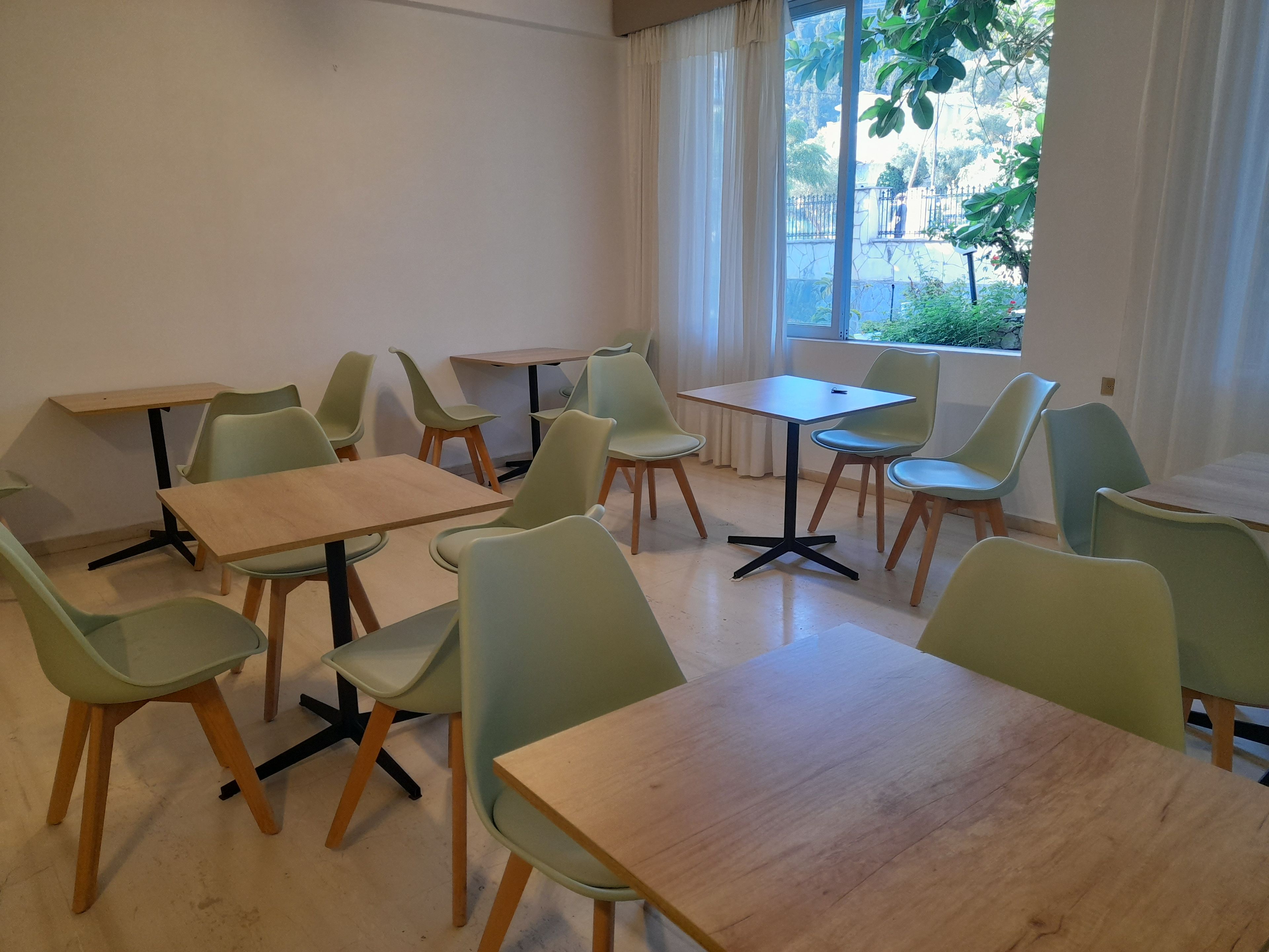 A room with multiple tables and chairs, large windows with curtains, and a view of greenery outside.