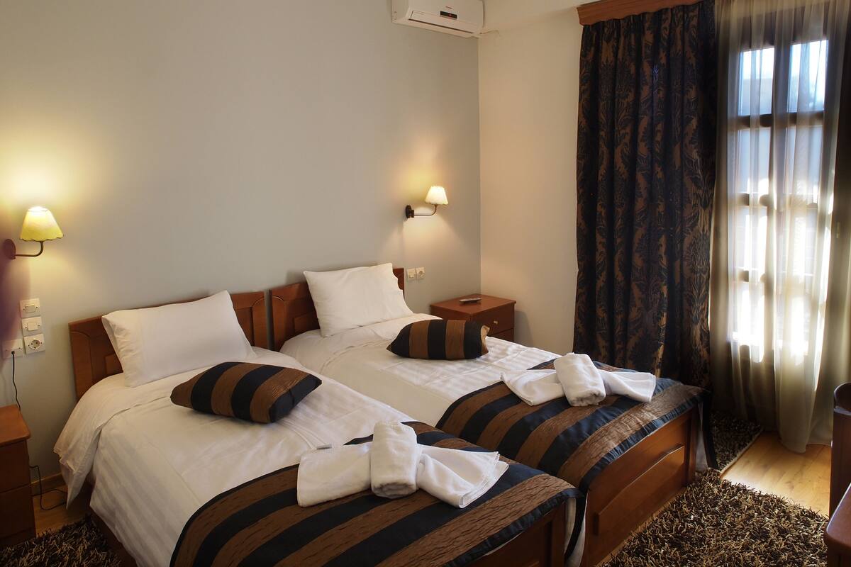 Hotelkamer met twee bedden, elk voorzien van gestreept beddengoed en kussens, een nachtkastje met een lamp, en een groot raam met gordijnen.