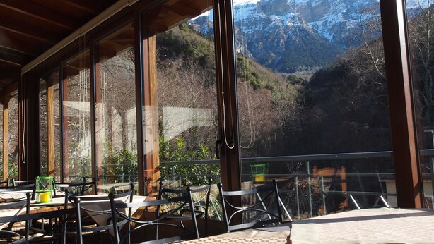 An outdoor dining area with tables and chairs, large windows offering a view of snow-capped mountains. - Archontiko Tzoumerkon (Central Tzoumerka)