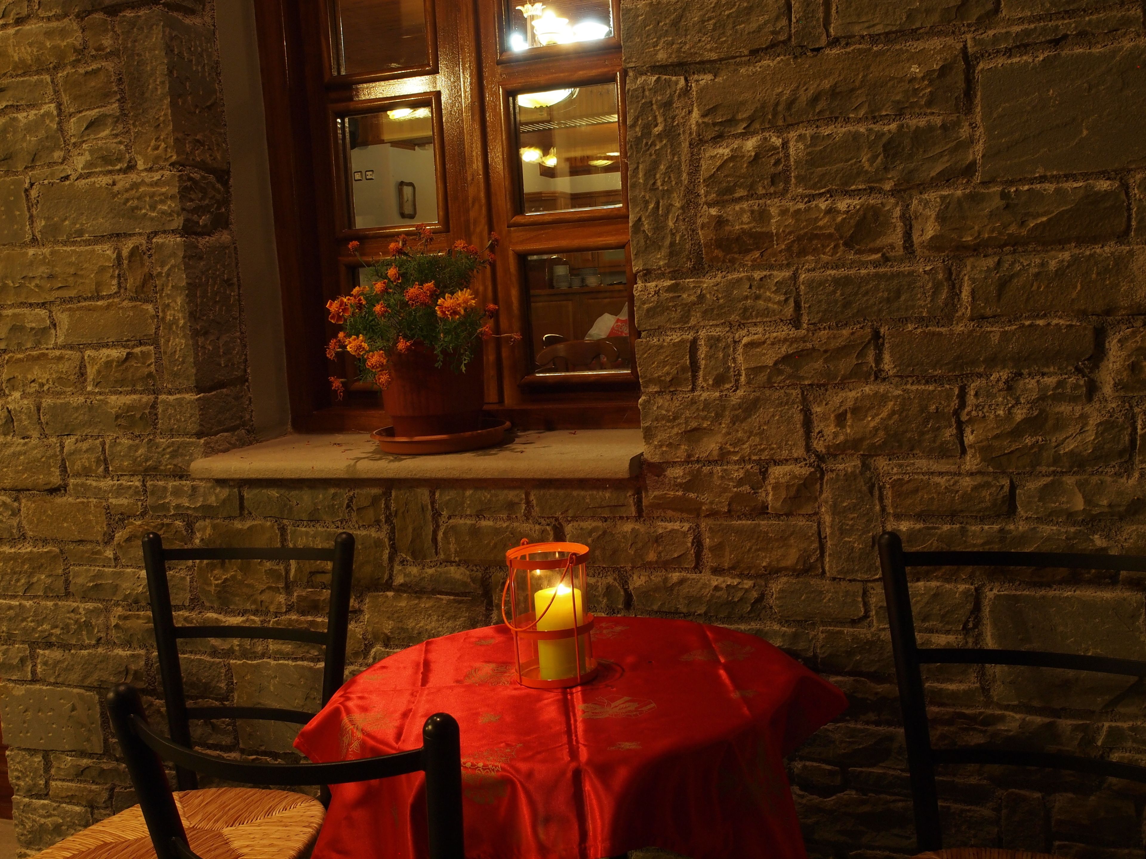 A stone wall with a window, a potted plant, a table with a red tablecloth, and two chairs.