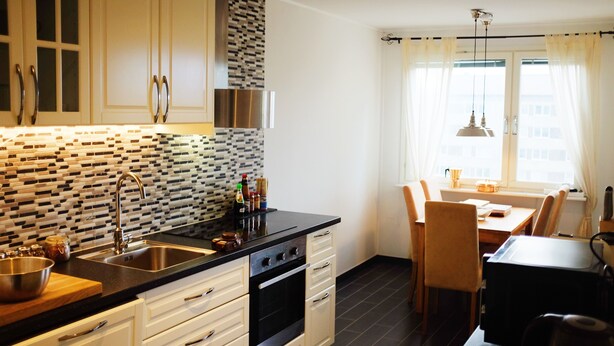 A modern kitchen with white cabinets, a black countertop, and a patterned backsplash. There is a dining area with a table and chairs, and a window with curtains. - Central - 2 Bedroom Apartment (Sollentuna)