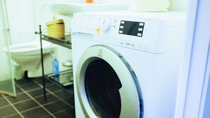 A front-loading washing machine with a digital display and control panel, situated in a bathroom with a tiled floor and a shelf above the toilet. - Central - 2 Bedroom Apartment (Sollentuna)