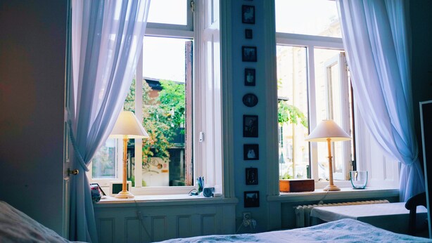 A bedroom with a bed, two bedside lamps, a window with curtains, a radiator, and a wall with framed pictures.