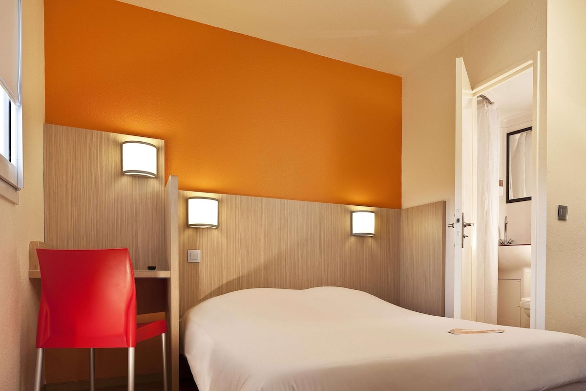 Une chambre d’hôtel avec un lit, un fauteuil rouge et deux lampes fixées au mur.