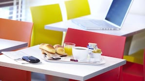Un plateau de petit-déjeuner avec du pain, du jus et du yaourt, posé sur une table dans une pièce aux couleurs vives, avec un ordinateur portable en arrière-plan.