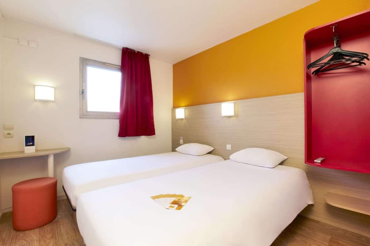 Une chambre d’hôtel avec deux lits, un mur rouge, une tête de lit en bois, une petite table et un tabouret rouge.