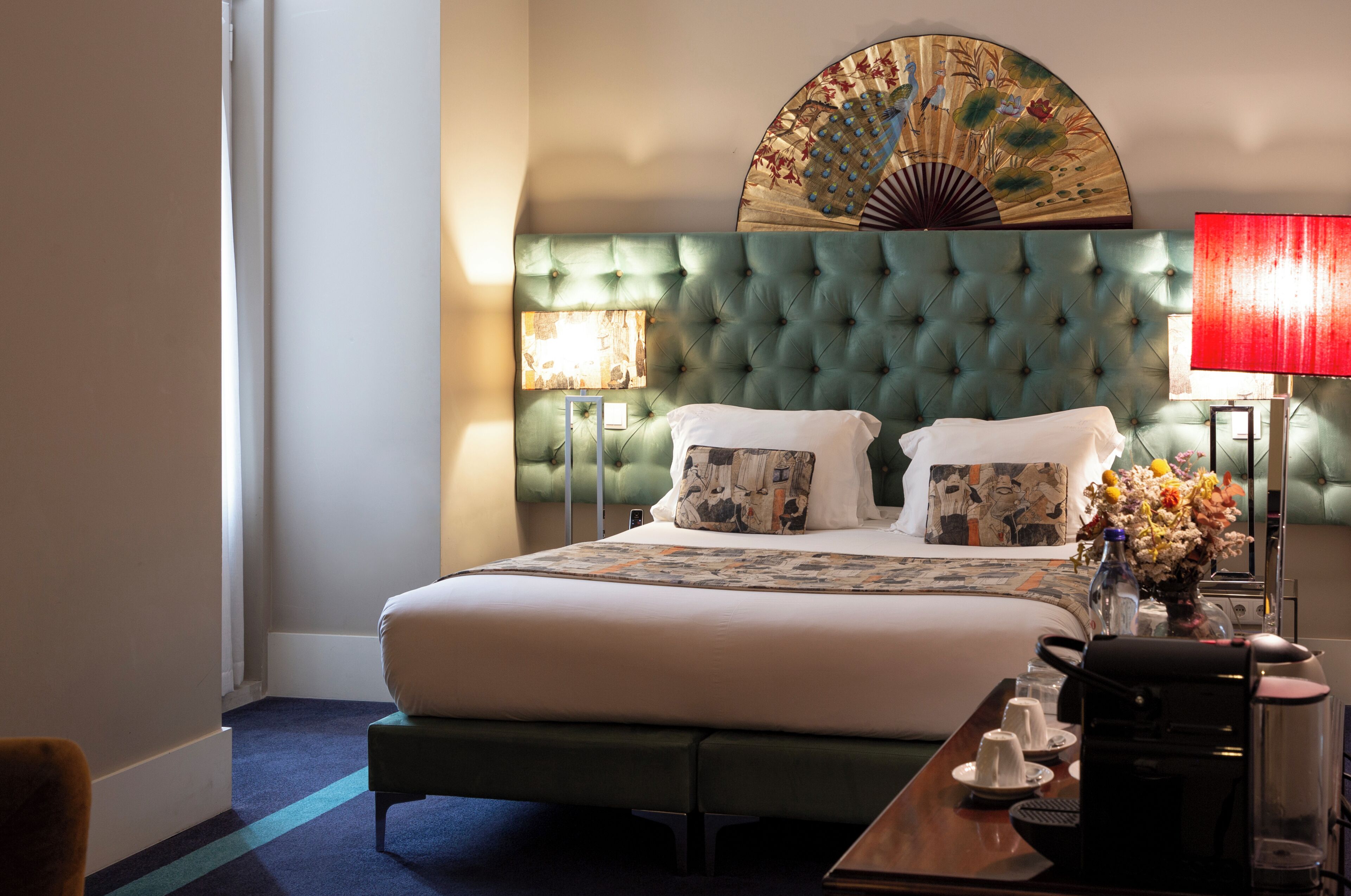 Une chambre d’hôtel avec une tête de lit capitonnée, un lit avec du linge de maison blanc, une table de chevet avec une lampe et un élément décoratif mural.