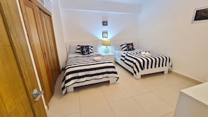 A bedroom with two beds, a wooden wardrobe, a nightstand with a lamp, and a small table. - Las Olas by Destinos 1A (Guayacanes)