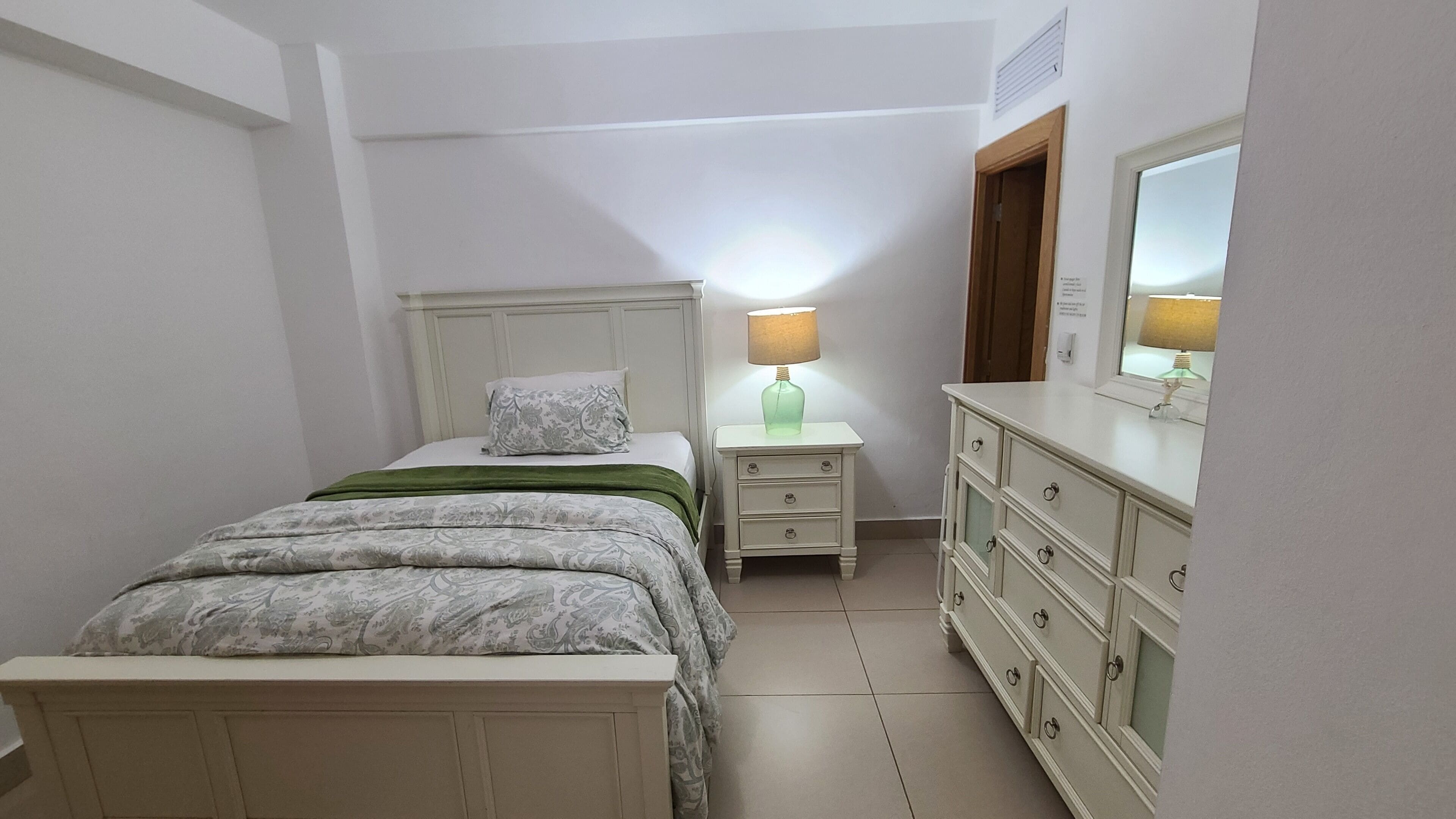 Un dormitorio con una cama, una mesita de noche con lámpara, un aparador y un espejo.
