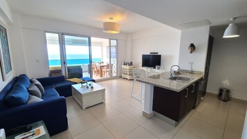 Una sala de estar moderna con sofá, una mesa de centro y televisor. Cuenta con una zona de cocina con pileta y una zona de comedor con sillas.